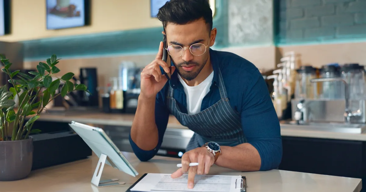 Cafe owner on the phone while checking a tablet and bills at the counter.