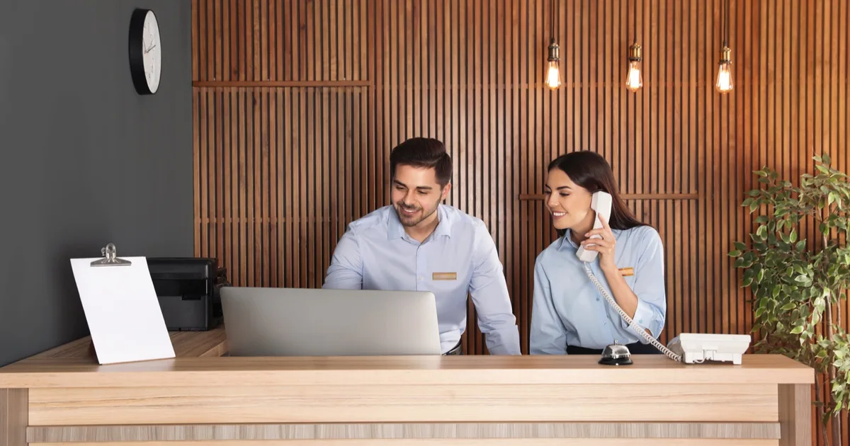 Hotel reception staff handling check-in with phone and laptop.